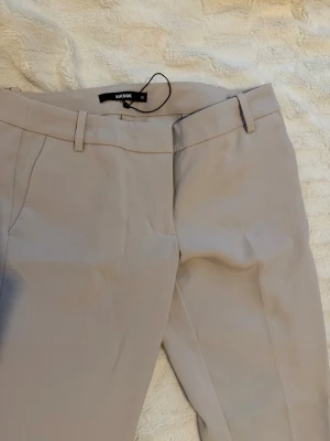 Lågmidjade kostymbyxor från Bik Bok - Snygga beige kostymbyxor från Bik Bok med raka ben och pressveck. Byxorna har hällor för skärp, sidofickor och en bakficka med minimalistisk design. Aldrig använt 