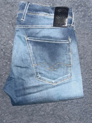 Blå slitna jeans från Replay - Säljer ett par blå jeans från Replay med slitningar och råa benslut. Jeansen har klassisk femficksdesign, ljus tvätt och dekorativa sömmar på bakfickan. Perfekta för en avslappnad och trendig look.