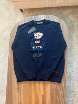 Mörkblå stickad tröja Polo Bear - Mörkblå stickad tröja från Polo Ralph Lauren med klassisk Polo Bear-design framtill.  • Storlek: M • Passform: Regular fit • Skick: Oanvänd – nyskick, med originaltag kvar • Färg: Mörkblå • Detaljer: Rund halsringning, ribbade muddar och nederkant, broderad Polo Bear med amerikansk flagga samt diskret “RL” på ärmen  Tröjan är helt oanvänd och i perfekt skick. En ikonisk modell från Ralph Lauren som är mycket eftertraktad och funkar lika bra till vardag som mer uppklätt.  Nypris: ca 2 500–3 000kr