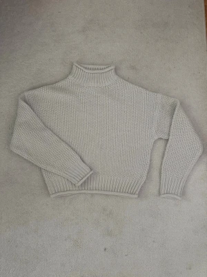 Ljusgrå stickad polotröja - Säljer en ljusgrå stickad polotröja med grov struktur och ribbade muddar vid ärmslut, nederkant och hals. Tröjan har en avslappnad passform och är perfekt för kyliga dagar. Materialet känns mjukt och mysigt.