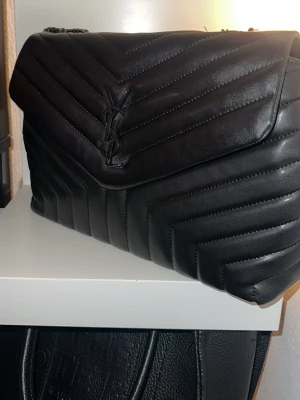 Svart YSL handväska i läder - Säljer en svart handväska med klassisk YSL-logga framtill. Väskan är tillverkad i quiltat läder med chevronmönster och har en kedjerem i silverfärg. Perfekt för dig som vill ha en lyxig och tidlös accessoar.