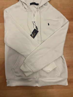 Vit hoodie från Polo Ralph Lauren - Snygg vit hoodie från Polo Ralph Lauren med dragkedja framtill och klassisk huva. Broderad logga i svart på bröstet och två stora fickor fram. Tillverkad i mjukt bomullsmaterial som känns skönt mot huden. Perfekt för en avslappnad och stilren look.