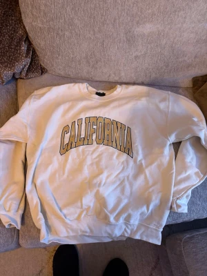 Vit sweatshirt med CALIFORNIA-tryck - Säljer en vit sweatshirt med stort guldfärgat CALIFORNIA-tryck över bröstet. Tröjan har rund halsringning, ribbade muddar vid ärmslut och nederkant samt en avslappnad passform. Perfekt för en chill och sportig look.