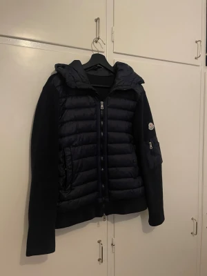 Svart pufferjacka från Moncler - Snygg svart pufferjacka från Moncler med stickade ärmar och vadderad front. Jackan har huva, dragkedja framtill och en ficka med Moncler-logga på ärmen. Perfekt för kalla dagar och riktigt trendig look.