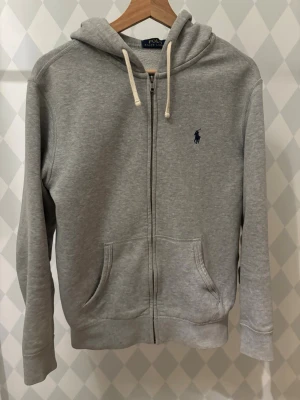 Grå hoodie från Polo Ralph Lauren - Snygg grå zip-hoodie från Polo Ralph Lauren med klassisk blå logga på bröstet. Hoodien har huva med vita snören, dragkedja framtill och två fickor. Perfekt för en avslappnad och stilren look.