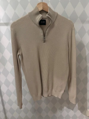 Beige stickad tröja Only & Sons - Snygg beige stickad tröja från Only & Sons med halv dragkedja och hög krage. Tröjan har ribbad struktur, långa ärmar och en avslappnad passform. Perfekt för lager på lager och enkel att matcha med olika outfits.
