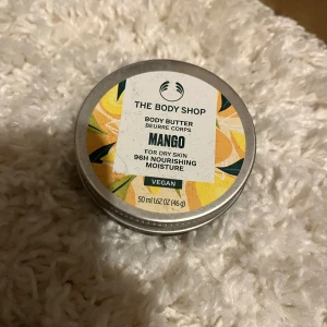 Mango Body Butter från The Body Shop - Mango Body Butter från The Body Shop i en rund metallburk med färgglad etikett. Innehåller 50 ml vegansk kroppssmörj för torr hud, ger fukt i upp till 96 timmar. Krämig konsistens med fruktig mangodoft.