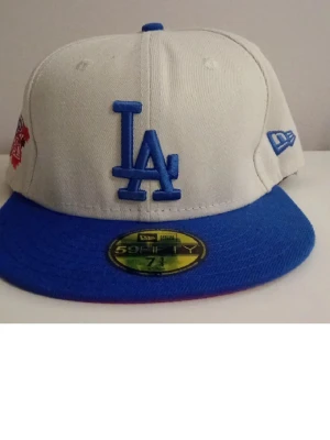 LA Dodgers keps från New Era - Snygg LA Dodgers keps från New Era 59FIFTY-serien. Kepsen har vit krona, blått broderat LA-logotyp framtill och blå skärm. Klassisk flat brim och broderad New Era-logga på sidan. Perfekt för dig som gillar streetwear och sportig stil.