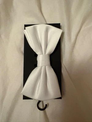 Vit rosett i satin - Snygg vit rosett i satinmaterial, perfekt som accessoar till olika outfits. Rosetten har en klassisk form och fästs enkelt. Passar dig som vill addera en stilren detalj till din look.