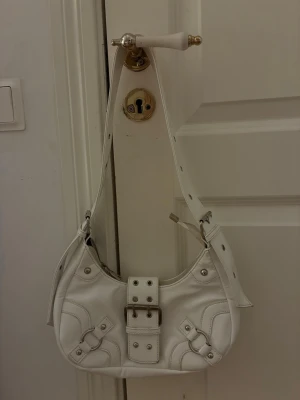 Vit axelremsväska med spänne, har använt den bara några gånger 🫶🏼 - Snygg vit handväska i skinnimitation med justerbar axelrem och stort dekorativt spänne framtill. Väskan har silverfärgade nitar och detaljer samt coola sömmar som ger en trendig look. Perfekt accessoar för att lyfta din outfit.