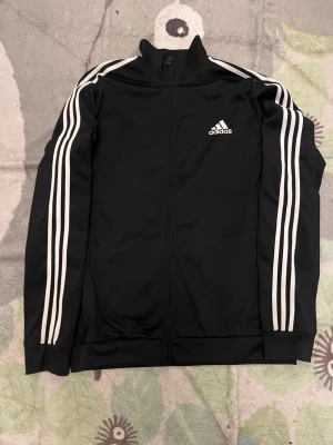 Svart Adidas träningsjacka - Svart träningsjacka från Adidas med klassiska vita ränder längs ärmarna och vit Adidas-logga på bröstet. Jackan har hel dragkedja, hög krage och muddar vid ärmslut och nederkant. Perfekt för sport eller chill. Materialet är troligtvis polyester.