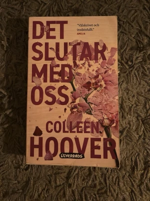 Det slutar med oss av Colleen Hoover - En gripande roman av Colleen Hoover med omslag i beige och lila toner, dekorerat med blommor. Boken handlar om Lily som kämpar med kärlek, relationer och sitt förflutna i Boston. Perfekt för dig som gillar känslosamma och insiktsfulla berättelser.