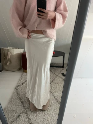 Vit långkjol i satin  - Säljer en elegant vit långkjol med slät och glansig satin-look. Kjolen har en rak och flowy passform som ger en stilren siluett. Perfekt att styla med en stickad tröja eller crop top för en trendig look.