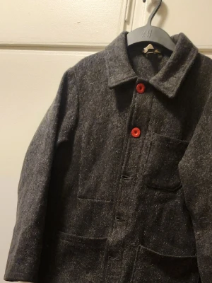 Le Laboureur mörkgrå ullkappa med stora fickor - Le Laboureur             Stilren mörkgrå ullkappa med raka linjer och stora utanpåliggande fickor framtill. Kappan har dold knäppning och är tillverkad i ett tjockt, värmande ullmaterial som passar perfekt för kyliga dagar. NY PRIS 2400KR 