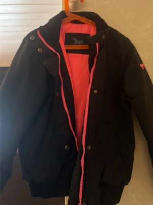 Svart bomberjacka med rosa detaljer till barn - Snygg svart bomberjacka med rosa foder och dragkedja. Jackan har ribbade muddar vid ärmslut och nederkant samt tryckknappar framtill. Perfekt för dig som vill ha en sportig och trendig look. Inköpt från hööks och blivit använd i stall