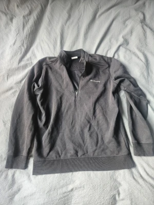 Svart halvzip från Columbia - Snygg mörkgrå tröja från Columbia med halv dragkedja framtill och diskret logga på bröstet. Tröjan har lång ärm och är tillverkad i ett mjukt material som passar perfekt till lager-på-lager. Enkel och stilren design.