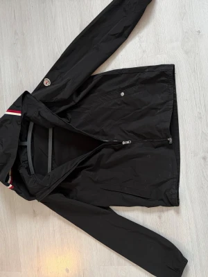 Svart vindjacka från Moncler - Snygg svart vindjacka från Moncler med huva och dragkedja framtill. Jackan har Moncler-logga på ärmen och detaljer i rött och vitt på huvan. Tillverkad i lätt och slitstarkt material, perfekt för blåsiga dagar. Fickor med dragkedja och knapp för extra funktion. Storlek L, ena knappen till fickan har låsnat där av pris går ner lite . Pris går att diskutera!