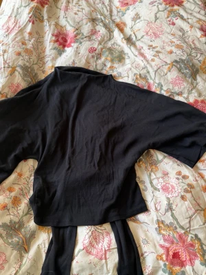 Svart croppad topp med knyt - Snygg svart croppad topp med korta ärmar och knyt i midjan. Toppen har en öppen rygg som knyts baktill för en trendig look. Perfekt att styla med höga jeans eller kjol.