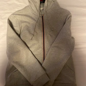 Grå hoodiejacka med dragkedja från Moncler - Grå hoodiejacka från Moncler med dragkedja framtill och huva med dragsko. Ränder i rött, vitt och blått på ärmslut och huva. Klassisk logotyp på bröstet. Perfekt för en sportig och stilren look.