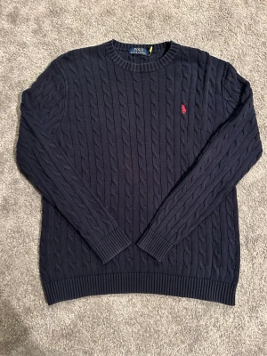 Mörkblå kabelstickad tröja Polo Ralph Lauren - Kabelstickad mörkblå tröja från Polo Ralph Lauren med rund halsringning och röd broderad logga på bröstet. Långa ärmar och ribbade muddar ger en klassisk look. Perfekt för lager-på-lager-stil.