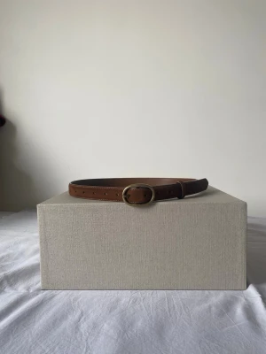 Brunt läderbälte med oval mässingsspänne - Snyggt brunt bälte i äkta läder med ett ovalt mässingsfärgat spänne. Bältet har flera hål för justering och en klassisk, stilren design som passar till jeans eller byxor. Perfekt accessoar för att lyfta din outfit.