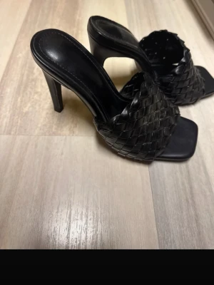 Svarta klackar - Snygga svarta klackar med fyrkantig tå och bred, flätad rem över foten. Skorna har en stabil klack och är tillverkade i ett material som ser ut som läder. Perfekta för dig som gillar trendiga och stilrena skor.