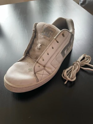 Vita DC sneakers i läder - Klassiska vita sneakers från DC med grå detaljer och stor logga på sidan. Skorna är låga och har en ovandel i läder samt en slitstark gummisula. Skosnören medföljer. Perfekta för dig som gillar streetwear och skate-inspirerad stil. Går även att be oss köpa nya skosnören innan köp av skorna. Pris kan duskuteras.