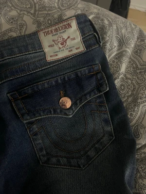 True Religion Joey Low Rise Flare Jeans - Säljer ett par mörkblå True Religion Joey jeans med låg midja och flare-ben. Klassisk kontrastsöm, ikonisk fickdesign och kopparfärgade knappar. Tillverkade i denim med snygg tvätt och detaljer som sticker ut.