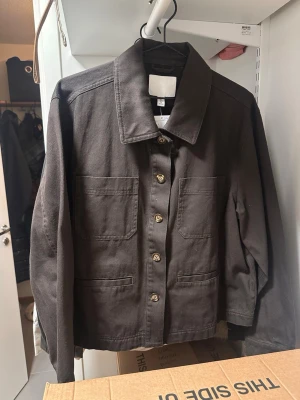 Mörkbrun overshirt från H&M - Stilren mörkbrun overshirt från H&M i kraftigt bomullstyg. Modellen har stora bröstfickor, klassisk krage och knäppning med träknappar framtill. 