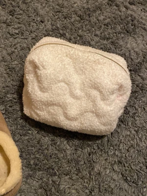 Vit fluffig necessär med dragkedja - Söt och mjuk necessär i vitt teddy-material med vågig struktur. Väskan har en guldfärgad dragkedja upptill och är perfekt för att förvara smink, småprylar eller andra viktiga saker. Enkel och stilren design som känns både trendig och mysig.