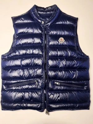 Moncler väst  - Snygg mörkblå dunväst från Moncler med glansig finish och klassisk logga på bröstet. Västen har två dragkedjeförsedda fickor framtill och hög krage med tryckknapp. Perfekt för lager-på-lager och riktigt street.