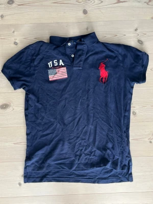 Marinblå piké från Polo Ralph Lauren - Säljer en marinblå pikétröja från Polo Ralph Lauren med klassisk krage, två knappar och stor röd logga på bröstet. USA-flagga och texten 'USA' broderat på vänster sida. Tillverkad i bomull och har normal passform.
