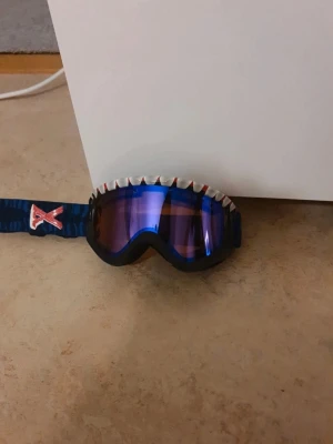 Blå skidglasögon  - Säljer ett par blå skidglasögon. Bredt blått band med röd och vit logga på sidan. Snygg accessoar för dig som vill sticka ut i backen. Glasögonen har en cool, sportig look och elastiskt band för bra passform.