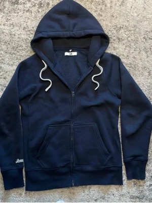 Mörkblå zip hoodie från Russell Russerch - Mörkblå hoodie med dragkedja från Russell. Klassisk modell med stor huva, vita dragsnören och två fickor framtill. Tillverkad i mjukt och tjockt material som känns riktigt skönt. Perfekt för dig som gillar en enkel och stilren look.