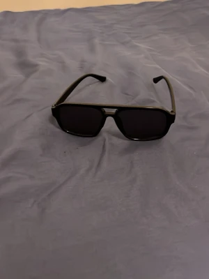 Svarta solglasögon med rektangulär form - Snygga svarta solglasögon med rektangulära glas och bred båge. Klassisk och stilren design som passar till de flesta outfits. Glasen är mörka och ramen är i plast för en lätt och bekväm känsla.