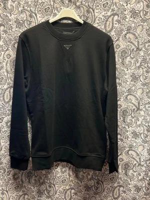 Svart sweatshirt från Prada - Stilren svart sweatshirt från Prada med diskret logga framtill vid halsen. Tröjan har rund halsringning, långa ärmar och ribbade muddar vid ärmslut och nederkant. Perfekt för en clean och modern look.
