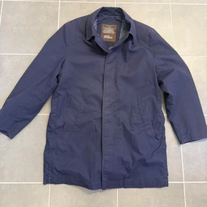 Mörkblå rock från Oscar Jacobsson  - Stilren mörkblå rock från Oscar Jacobsson med klassisk krage och dold knappslå. Rocken har två sidofickor och en rak passform som ger en clean look. Perfekt för dig som vill ha en enkel men snygg jacka till vardags. Materialet är slätt och lätt, vilket gör den smidig att bära.