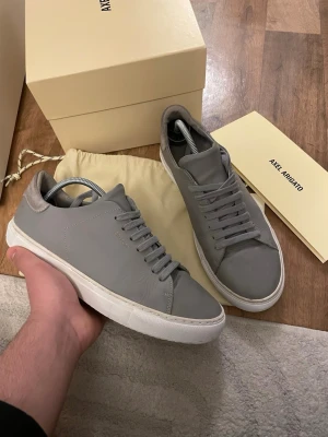 Gråa sneakers från Axel Arigato - Stilrena grå sneakers från Axel Arigato i slät läder med matchande grå skosnören och vit sula. Diskret logga i guld på sidan och klassisk låg profil. Perfekta för dig som gillar minimalistisk och clean stil. Box med följer ej 