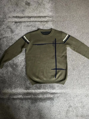 Olivgrön stickad tröja från strömskär - Säljer en olivgrön stickad tröja med rund hals och långa ärmar. Tröjan har vita och mörkblå ränder på axlarna samt ett grafiskt mörkblått mönster framtill. Perfekt för dig som gillar enkel men unik design. Material är premiumull