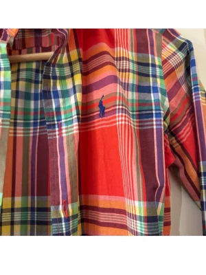 Rutig skjorta från Ralph Lauren Sport - Färgglad rutig skjorta från Ralph Lauren Sport med klassisk krage och knappar framtill. Skjortan har flera starka färger som röd, blå, grön och gul samt en broderad logga på bröstet. Tillverkad i mjuk bomull, perfekt för en avslappnad men stilsäker look.