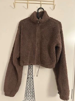 Brun cropped teddyjacka med dragkedja från NLY Trend  - Mysig brun teddyjacka i cropped modell med hög krage och dragkedja framtill från NLY Trend i storlek XS. Jackan har långa ärmar och justerbar dragsko i nederkant. 