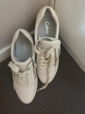 Vita sneakers från Gabor med dragkedja - Snygga vita sneakers från Gabor med klassisk låg profil. Skorna har både snörning och dragkedja på sidan för enkel på- och avtagning. Ovandelen är i slätt läder och sulan är i gummi. Perfekta för en clean och stilren look.