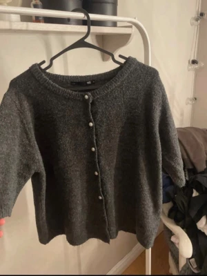 Mörkgrå kofta från lager - Mysig mörkgrå kofta från lager med rund halsringning och pärlknappar framtill. Koftan är stickad och har en rak, avslappnad passform samt långa ärmar. Perfekt att slänga över en t-shirt eller linne för extra värme.
