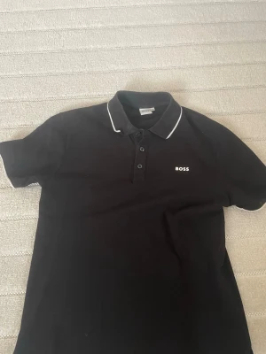 Svart pikétröja från BOSS - Klassisk svart pikétröja från BOSS med vit piping på kragen och liten BOSS-logga broderad på bröstet. Tröjan har korta ärmar, knappar framtill och är tillverkad i mjukt bomullsmaterial. Perfekt till en stilren och enkel look. (äkta hugo boss)