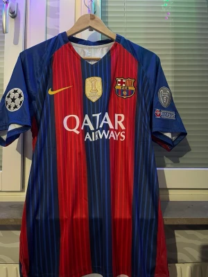 Barcelona matchtröja Nike blå/röd storlek L - Säljer en officiell FC Barcelona matchtröja från Nike med vertikala blå och röda ränder. Tröjan har Qatar Airways som sponsortryck, Champions League-märken på ärmarna och klubbmärke på bröstet. Tillverkad i lätt och ventilerande material, perfekt för fotboll.