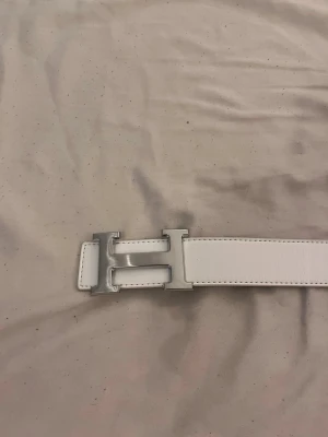 Vitt skärp med H-spänne - Snyggt vitt skärp med ett stort H-format spänne i silverfärg. Skärpet är gjort i slätt skinn och har svarta kanter på baksidan. Perfekt accessoar för att lyfta din outfit.