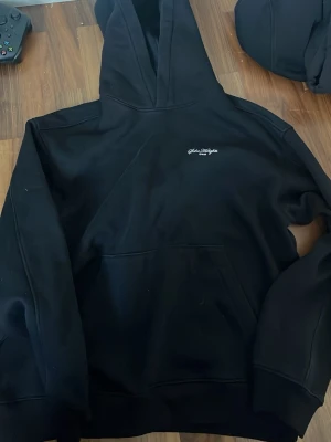 Svart hoodie från h&m - Snygg svart hoodie från H&M med liten vit logga på bröstet och stor, stilren SH-print i vitt på ryggen. Klassisk känguruficka framtill och stor huva. Tillverkad i mjukt bomullsmaterial som är perfekt för chill dagar.