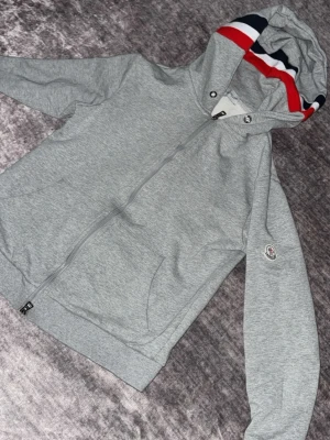 Storlek s Grå Moncler hoodie med dragkedja - Storlek s Snygg grå hoodie från Moncler med hel dragkedja och huva. Huven har coola ränder i rött, vitt och marinblått. Klassisk Moncler-logga på ärmen och kängurufickor framtill. Perfekt för en chill och sportig look.
