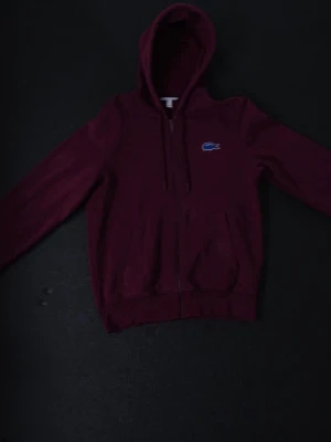 Lacoste Hoodie vinröd (S) - Snygg vinröd hoodie från Lacoste med dragkedja framtill och klassisk krokodil-logga på bröstet. Hoodien har huva med dragsko och två fickor framtill. Perfekt för en chill och stilren look. Tillverkad i mjukt bomullsmaterial.