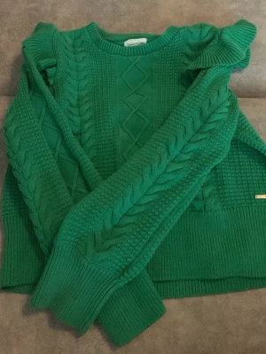 Grön stickad tröja från HOLLY & WHITE  - Snygg grön stickad tröja från Emily & White med flätstickat mönster på ärmar och framstycke. Tröjan har ribbade muddar, rund halsringning och dekorativa volanger vid axlarna. Perfekt för dig som vill ha något färgglatt och trendigt i garderoben.